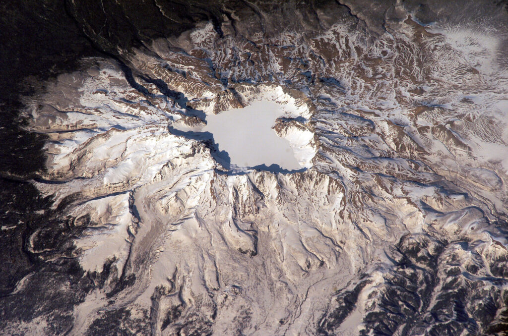 Snow-covered Baitoushan Volcano with frozen Lake Tianchi at its summit, showing caldera walls and lava flow patterns, as seen from the International Space Station.